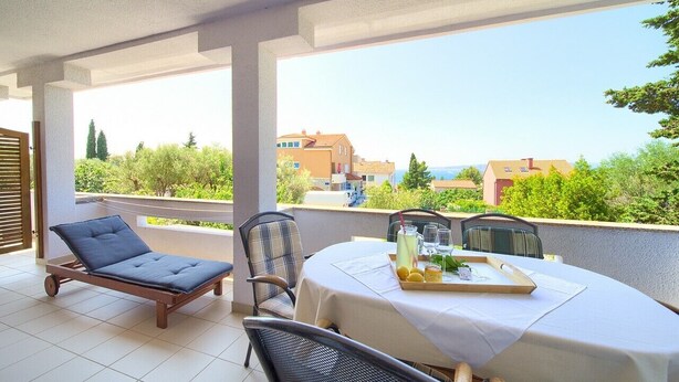 A balcony with a table set for dining, a lounge chair, and a view of buildings and greenery. - Apartments Loncar (Krk)