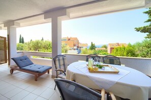 Un balcón con mesa lista para comer, una silla de lounge y vistas a edificios y vegetación.