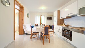 A modern kitchen with white cabinets, a built-in oven, and a dining area with wooden chairs and a table set for four. - Apartments Loncar (Krk)