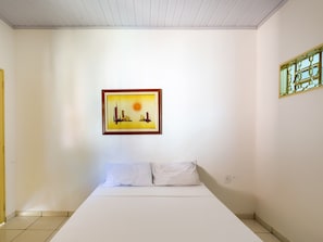 A bedroom with a bed, white walls, a framed picture of a cityscape, and a window with a decorative frame.