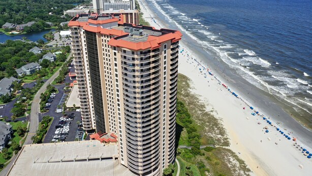 A high-rise hotel with a curved roof, adjacent to a beach with umbrellas and a parking lot. - OCEANVIEW 308 MARGATE - Bright Beach Decor, NEW FLOORS, & Ready to Accommodate!  (Myrtle Beach)