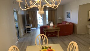 A dining area with a table set for two, a vase of flowers, and a red sofa in the background. - OCEANVIEW 308 MARGATE - Bright Beach Decor, NEW FLOORS, & Ready to Accommodate!  (Myrtle Beach)