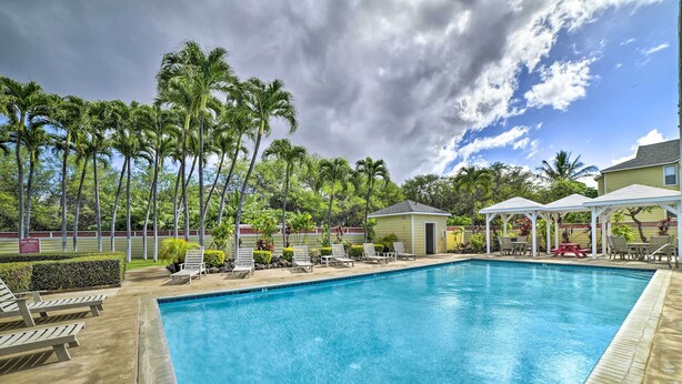 A swimming pool with lounge chairs and a pavilion, surrounded by palm trees and a cloudy sky.