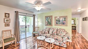 A living room with a floral sofa, wooden flooring, a ceiling fan, and a sliding glass door leading to a balcony.