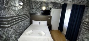 A hotel room with a marble-patterned wall, a bed with white linens, a nightstand, a desk, a chair, a desk lamp, and a window with curtains. - Zirkon Suit Otel (Istanbul)