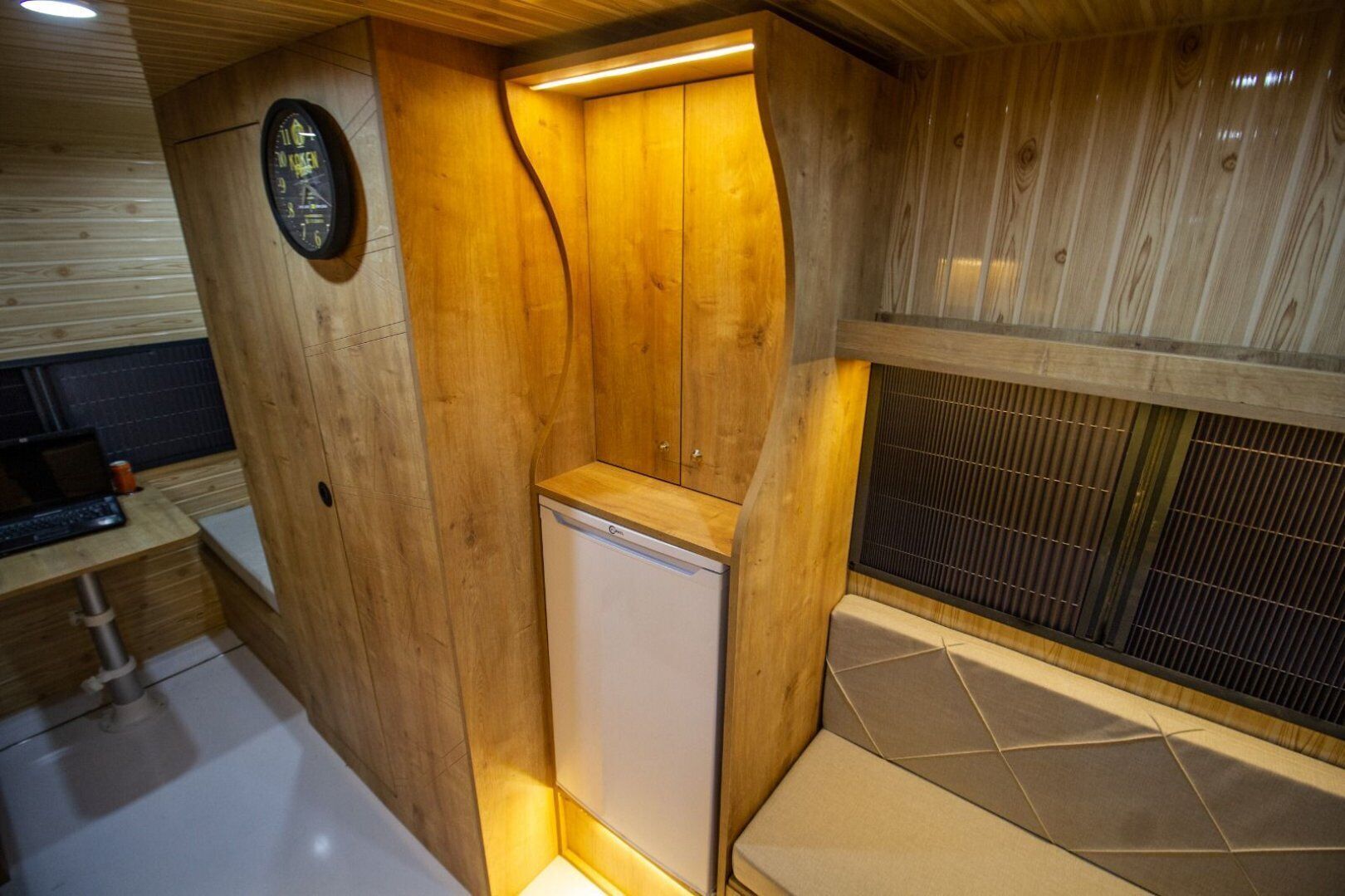 Un sauna compact, aux finitions en bois, doté d’un banc intégré et d’un petit appareil blanc, probablement un radiateur ou un humidificateur, placé dans un coin avec un sol carrelé et un mur en bois orné d’une horloge.