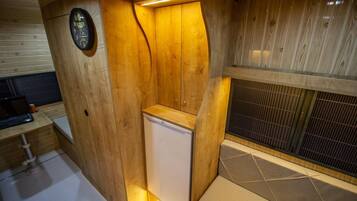 Un sauna compact, aux finitions en bois, doté d’un banc intégré et d’un petit appareil blanc, probablement un radiateur ou un humidificateur, placé dans un coin avec un sol carrelé et un mur en bois orné d’une horloge.