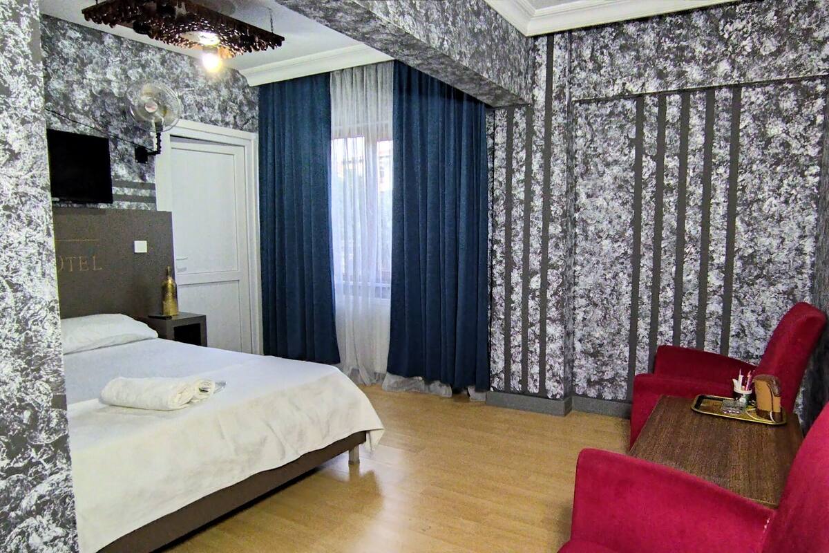 Habitación de hotel con cama, un sillón rojo, una mesita con bandeja y una ventana con cortinas azules.