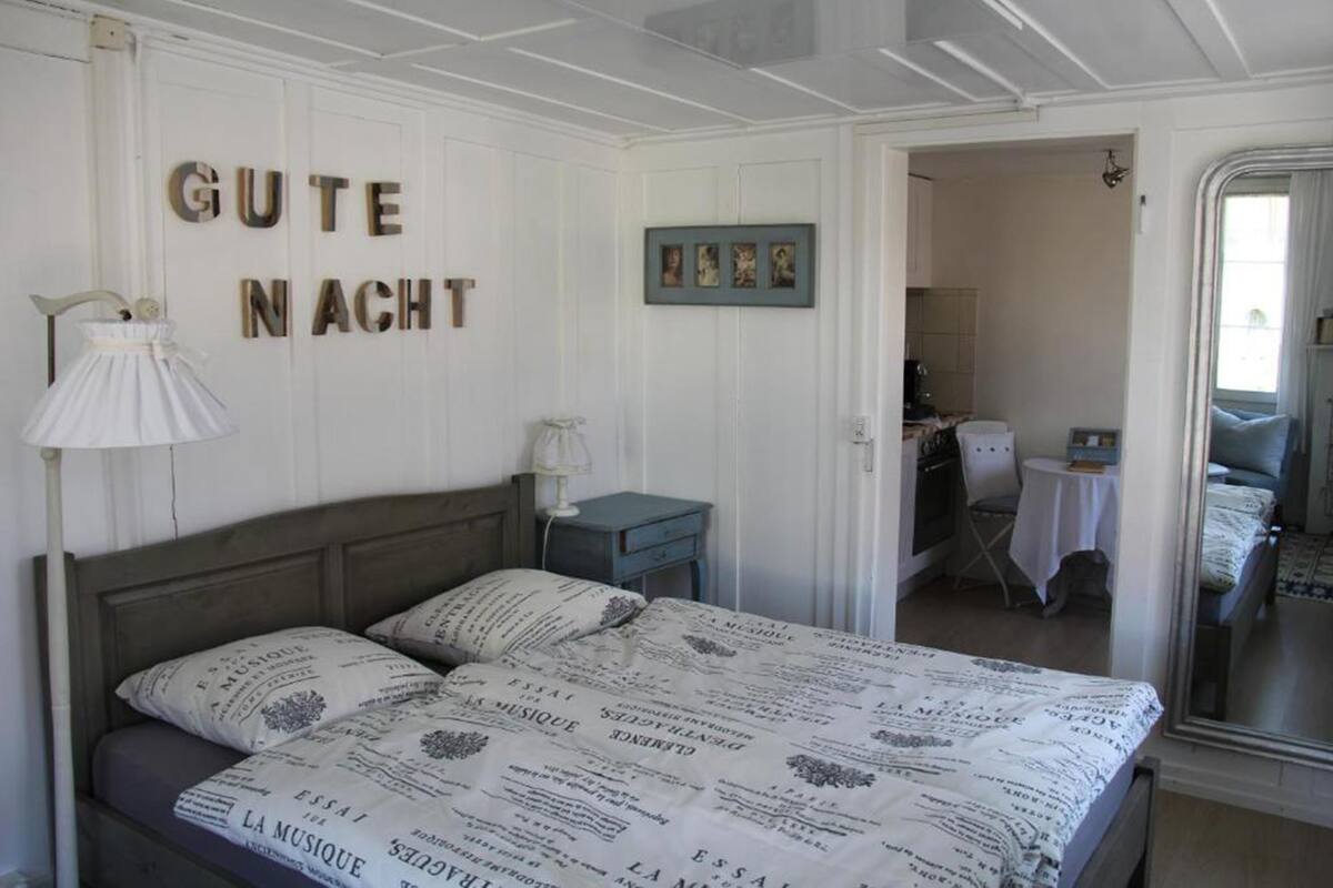 Ein Schlafzimmer mit einem Bett, einem Nachttisch, einer Lampe und einem Spiegel. An der Wand hängen Bilder in Rahmen, und eine Tür führt in einen anderen Raum.