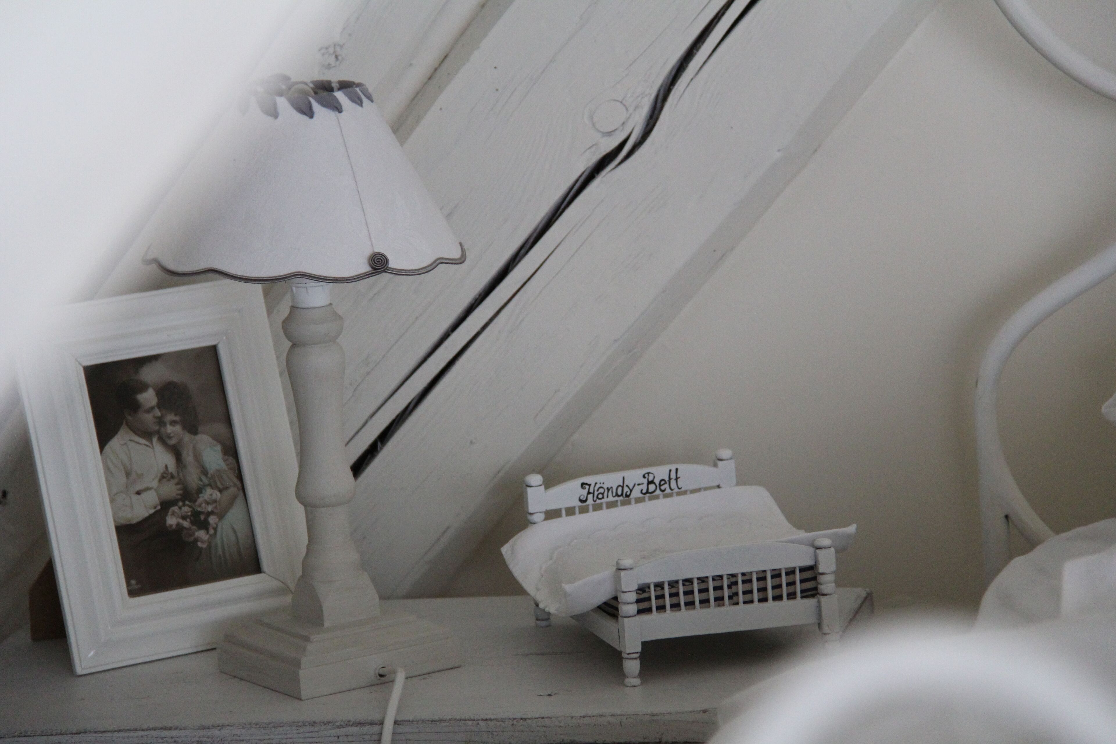 Una cama blanca pequeña con cabecera identificada con la leyenda “Handy-Bell”, una lámpara blanca y una fotografía en un marco sobre una superficie de madera.