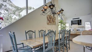 Un comedor con mesa de madera y sillas azules, una araña y vistas a la montaña a través de amplias ventanas.