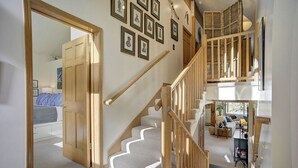 Una escalera con barandillas de madera que lleva a un segundo piso, un pasillo con cuadros enmarcados y una habitación con cama y ventana.