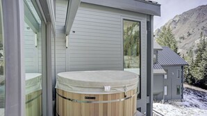 Una casa moderna con un jacuzzi de madera en el porche, un cobertizo pequeño y un paisaje montañoso nevado.