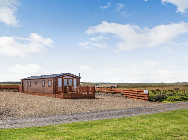 A wooden cabin with a fenced yard, a gravel path, and a grassy field in the background.