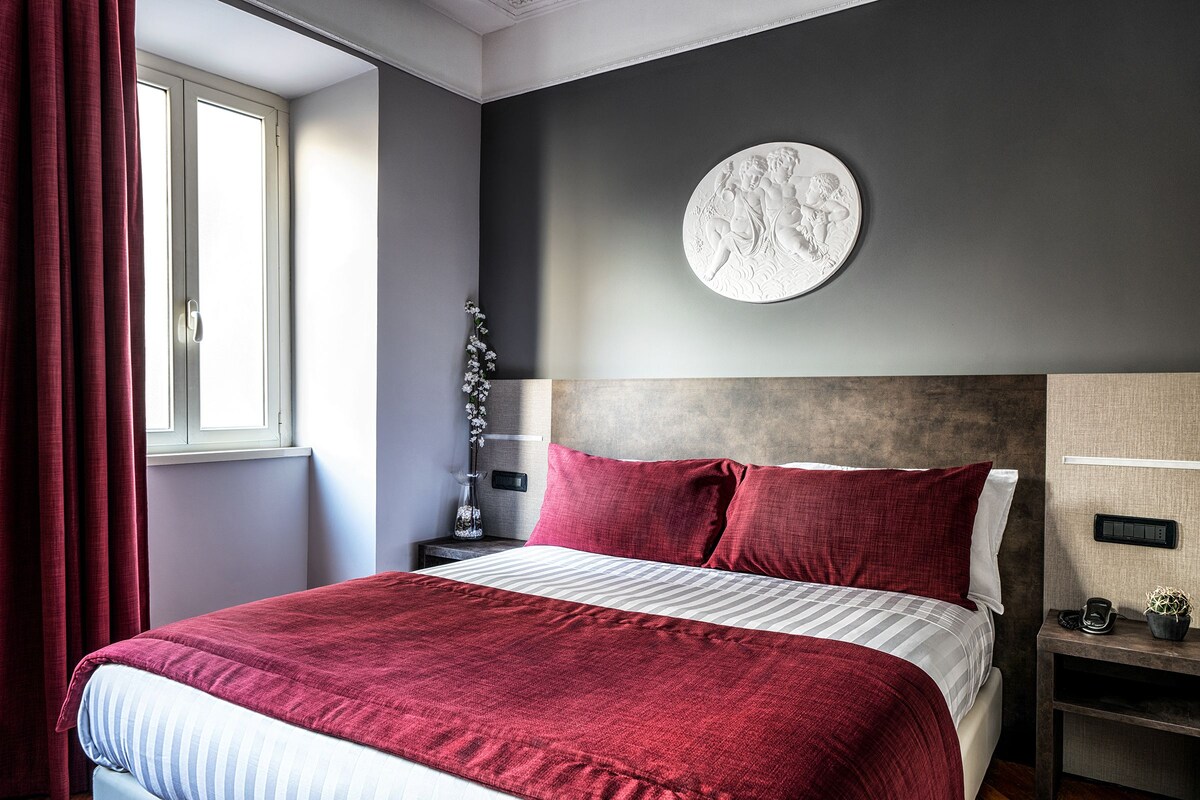 A modern bedroom with a large bed, red bedding, a round wall decoration, and a nightstand with a phone and a small plant.