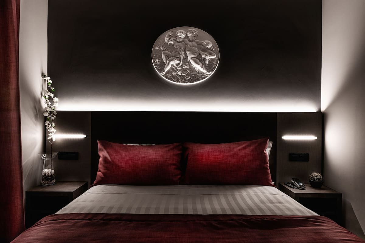 A modern bedroom with a bed, red pillows, a wall-mounted circular artwork, bedside tables with lamps, and a vase with flowers.