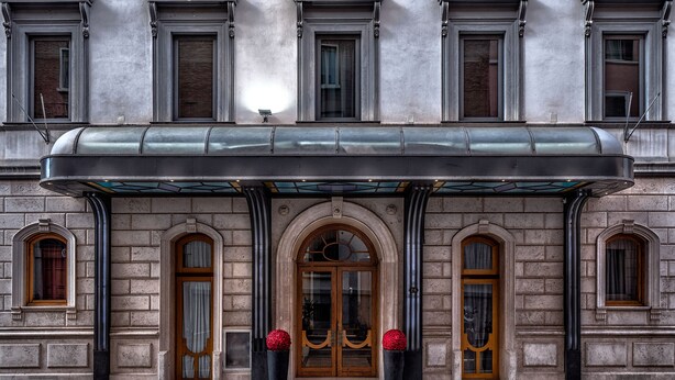 A building facade with a central arched entrance, flanked by two symmetrical windows with shutters, and a covered entrance area with a glass canopy. - Hotel Varese Roma (Rome)