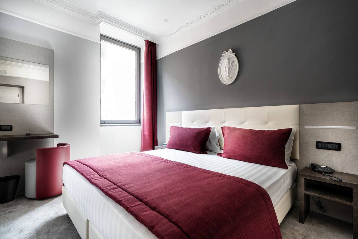 A modern hotel room with a large bed, a desk with a telephone, and a wall-mounted clock.