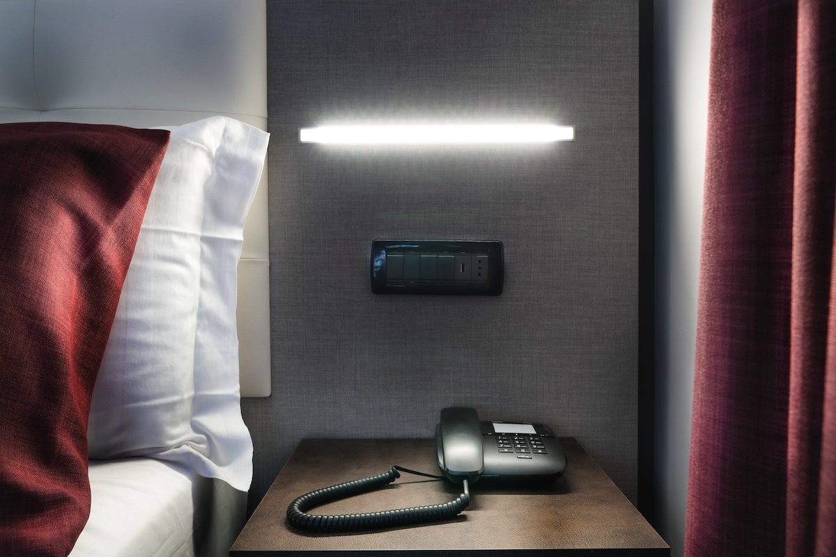 A hotel room with a bed, a bedside table with a phone, and a wall-mounted light.