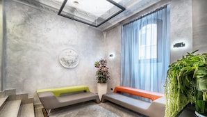 A modern waiting area with a marble floor, a green and orange bench, a potted plant, and a large window with curtains. - Hotel Varese Roma (Rome)