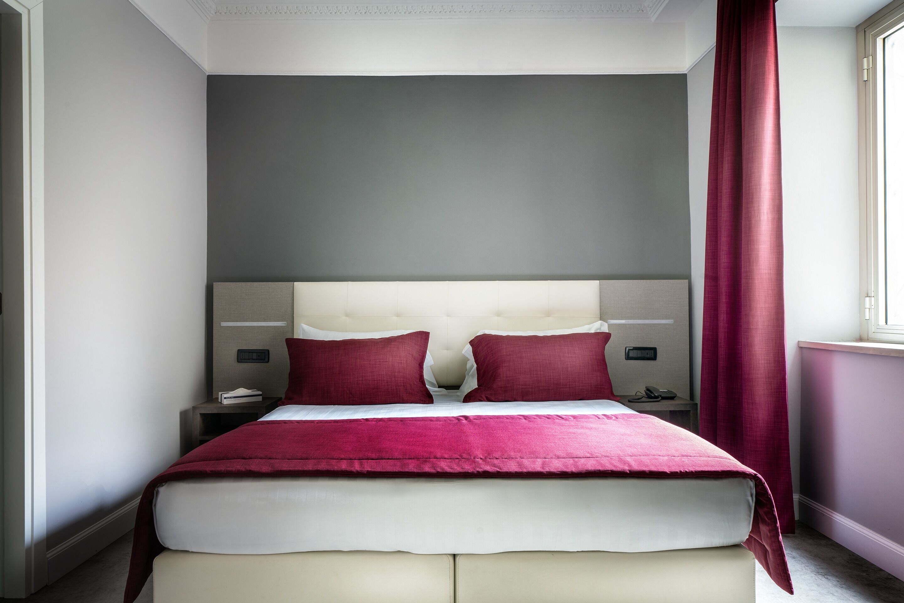 A neatly made bed with a red blanket and pillows, a bedside table with a lamp, and a window with red curtains.