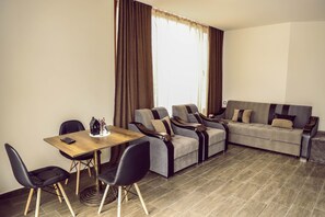 Ein modernes Wohnzimmer mit einer Couch, zwei Sesseln, einem Esstisch mit Stühlen und einem großen Fenster mit Vorhängen.