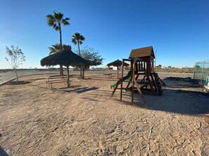 Ein Sandspielplatz mit einer Holzschaukel und einem strohgedeckten Gebäude.