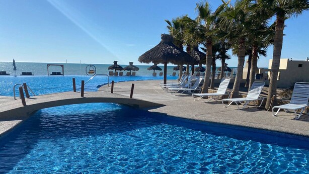 Une piscine à l’eau d’un bleu cristallin, entourée de chaises longues et de palmiers, avec une vue sur l’océan en arrière-plan.