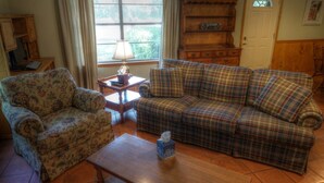 A living room with a floral armchair, a plaid sofa, a wooden side table with a lamp, and a bookshelf.