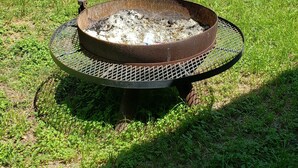 A metal fire pit with a wire grate, surrounded by grass and trees in a park setting.
