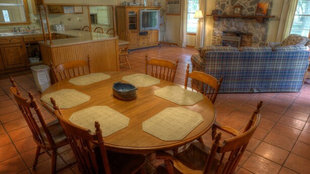 A dining area with a round wooden table surrounded by chairs, a kitchen in the background, and a blue plaid sofa in the living area.
