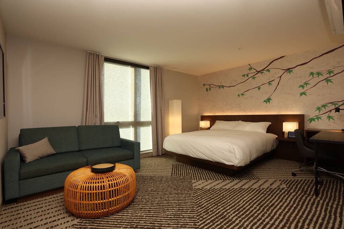 A hotel room with a bed, a sofa, a round wicker ottoman, a desk, and a wall with a branch design.