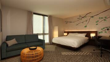 Habitación de hotel con cama, sofá, reposapiés redondo de mimbre, escritorio y una pared con diseño de ramas.