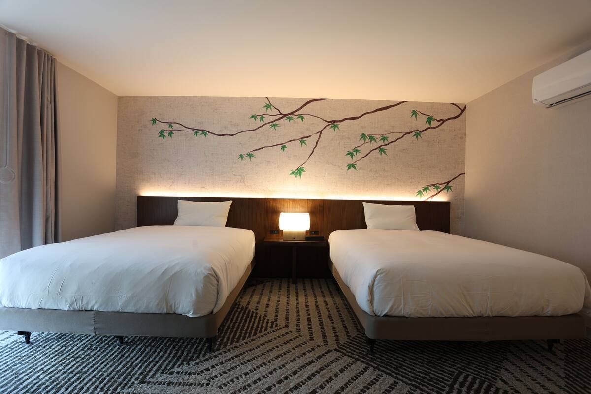 A hotel room with two beds, a nightstand with a lamp, and a wall mural of a tree branch.