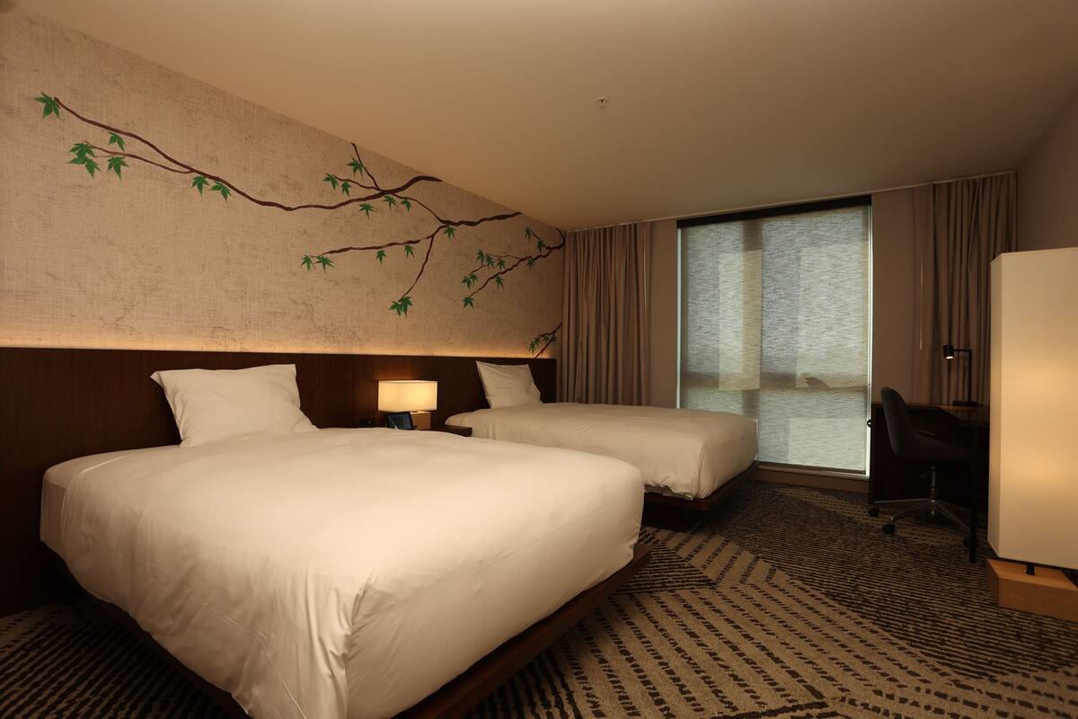 A hotel room with two beds, a desk, a chair, a large window, and a wall mural.