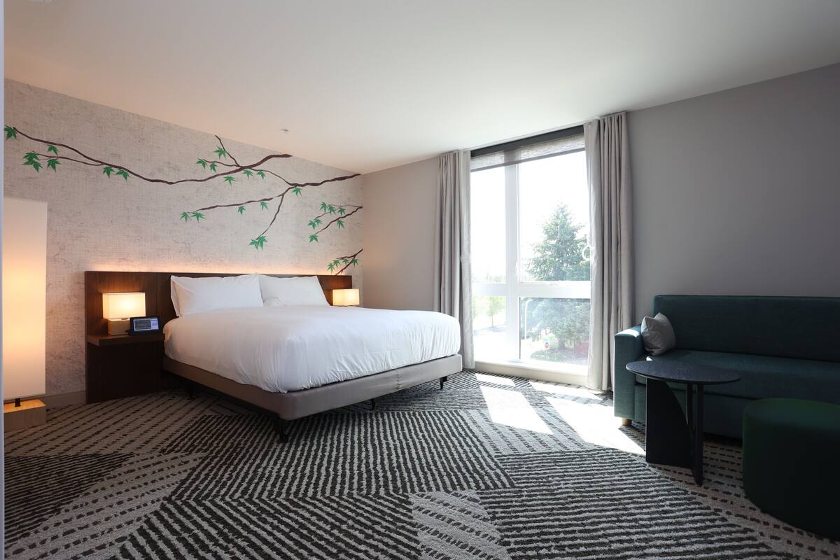 A modern hotel room with a large bed, a small sofa, a side table, and a view of the outdoors.