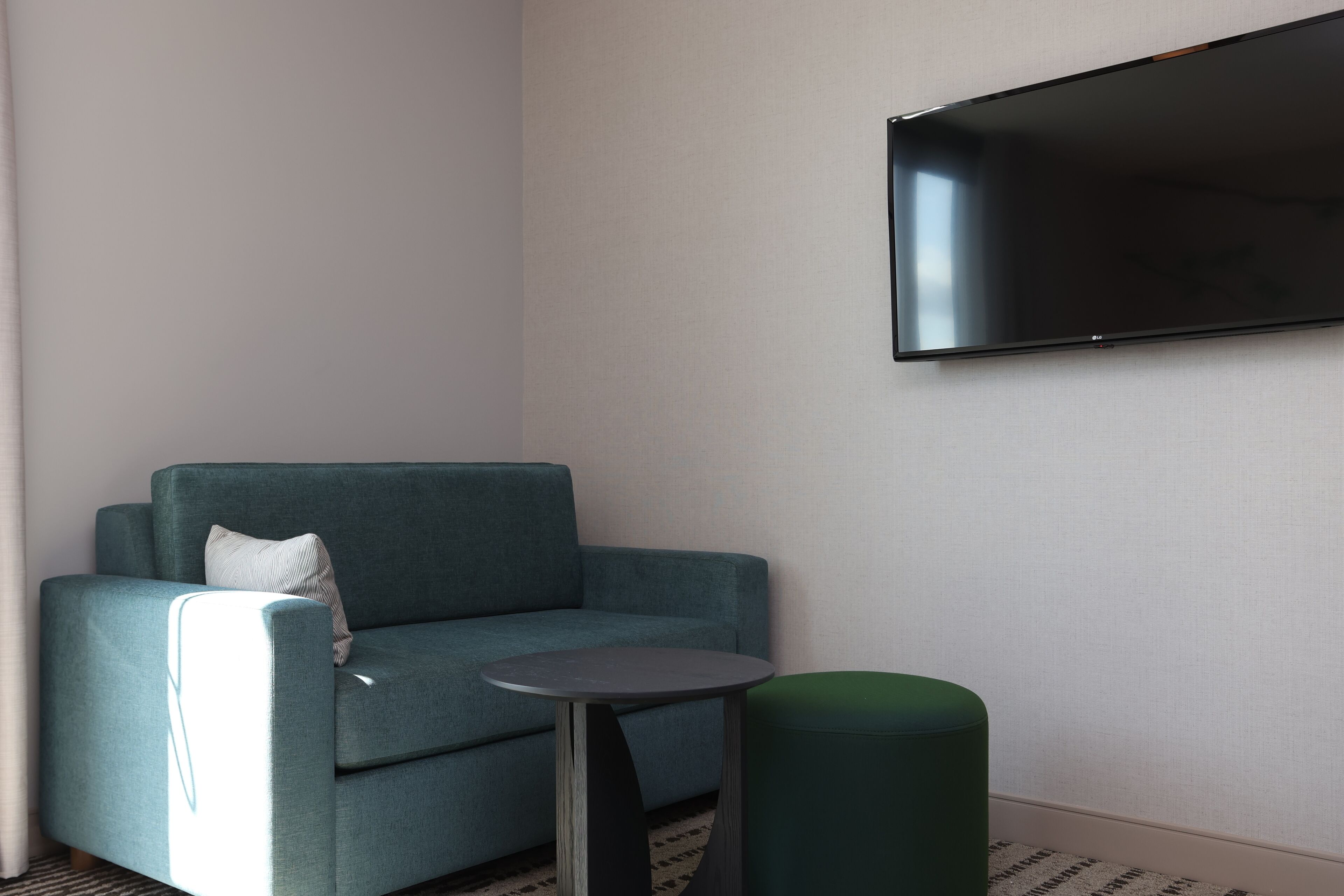 Un salon moderne avec un canapé de couleur turquoise, une petite table ronde et un pouf vert. Un téléviseur à écran plat est fixé au mur.