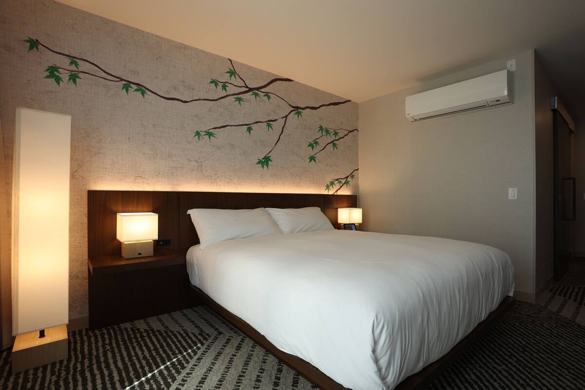 A hotel room with a large bed, two bedside lamps, a wall-mounted air conditioner, and a patterned rug.