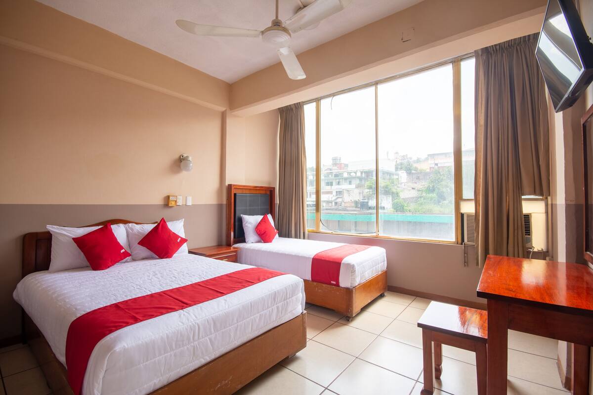 A hotel room with two beds, a desk, a ceiling fan, and a large window with curtains.