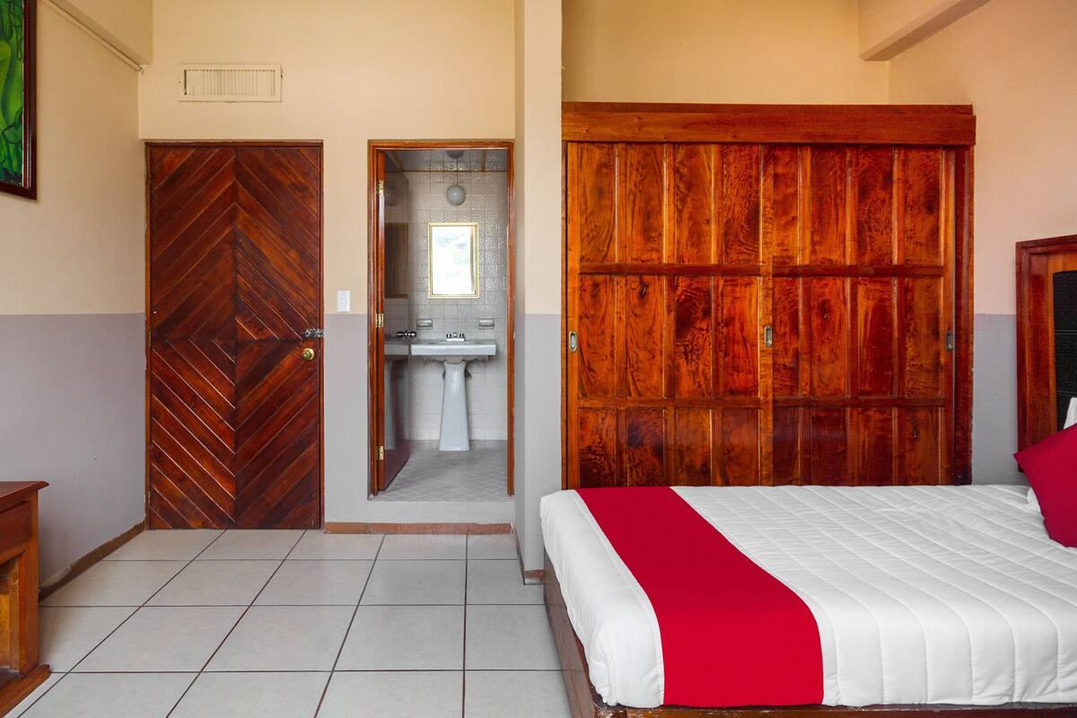 A bedroom with a bed, a wooden wardrobe, a bathroom with a sink, and a door leading to another room.