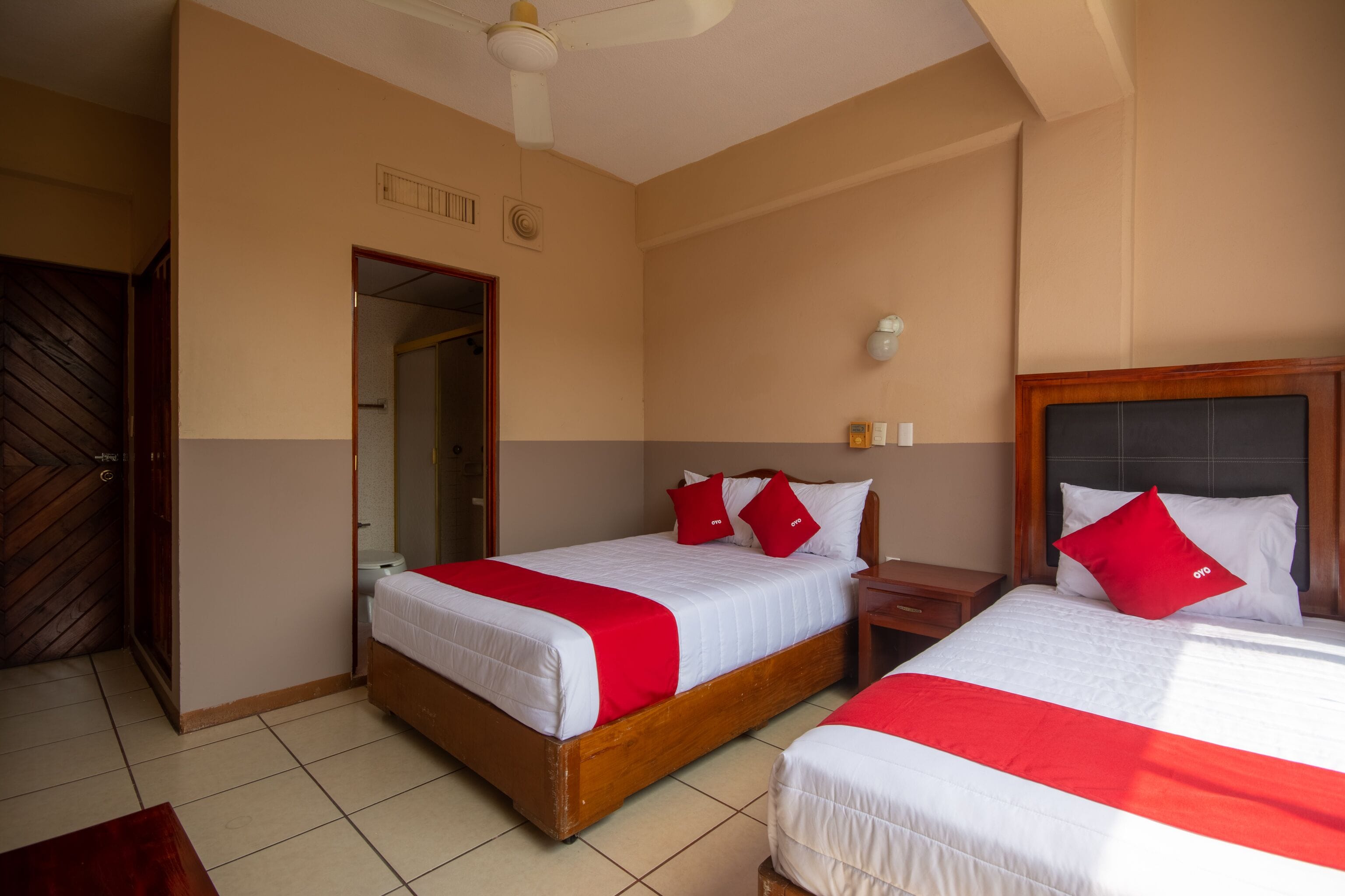 Habitación de hotel con dos camas, una puerta de madera, un ventilador de techo y piso de baldosas.