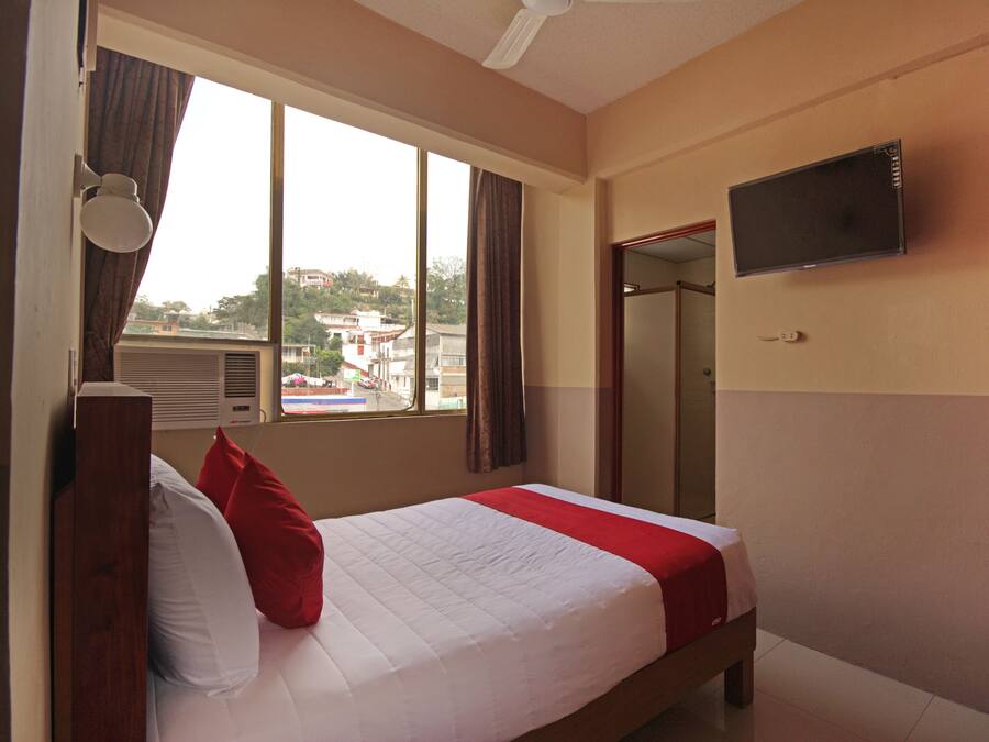 A hotel room with a bed, a TV mounted on the wall, a window with curtains, and a view of the outside.
