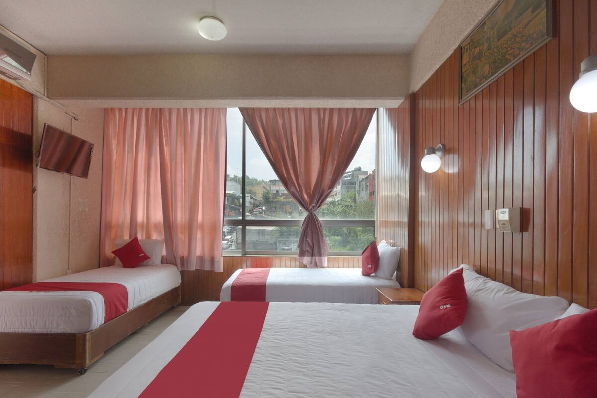 A hotel room with two single beds, a larger bed, wooden paneling, a view of the city, and a mounted television.