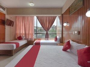 Habitación de hotel con dos camas individuales, una cama más grande, paneles de madera, vista a la ciudad y un televisor montado.