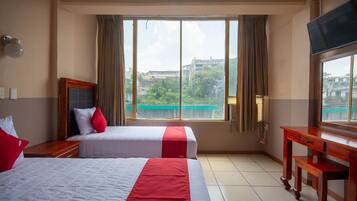 A hotel room with two beds, a desk, a TV, and a large window with curtains.
