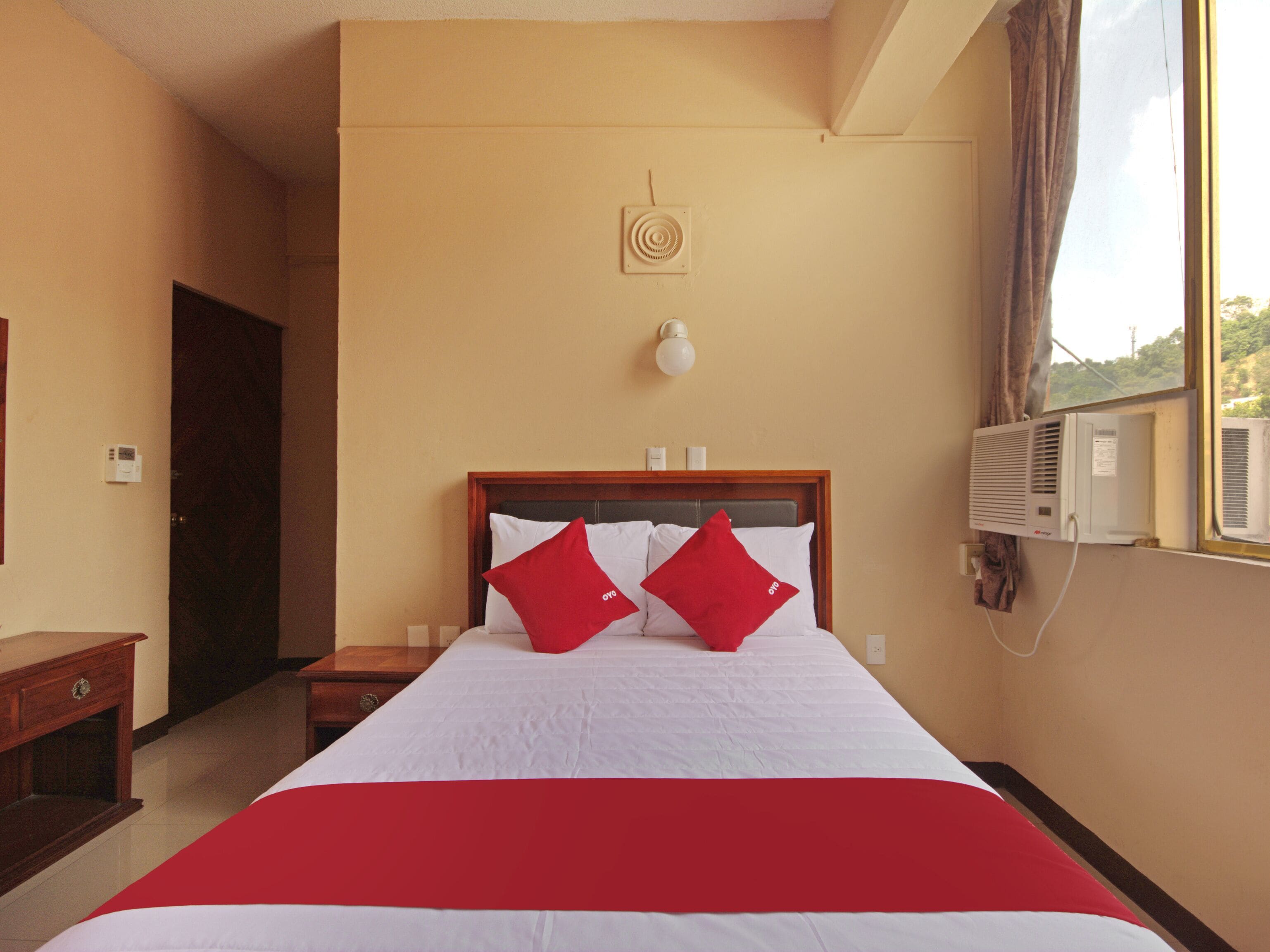 A hotel room with a bed, a bedside table, a wall-mounted air conditioner, and a window with curtains.