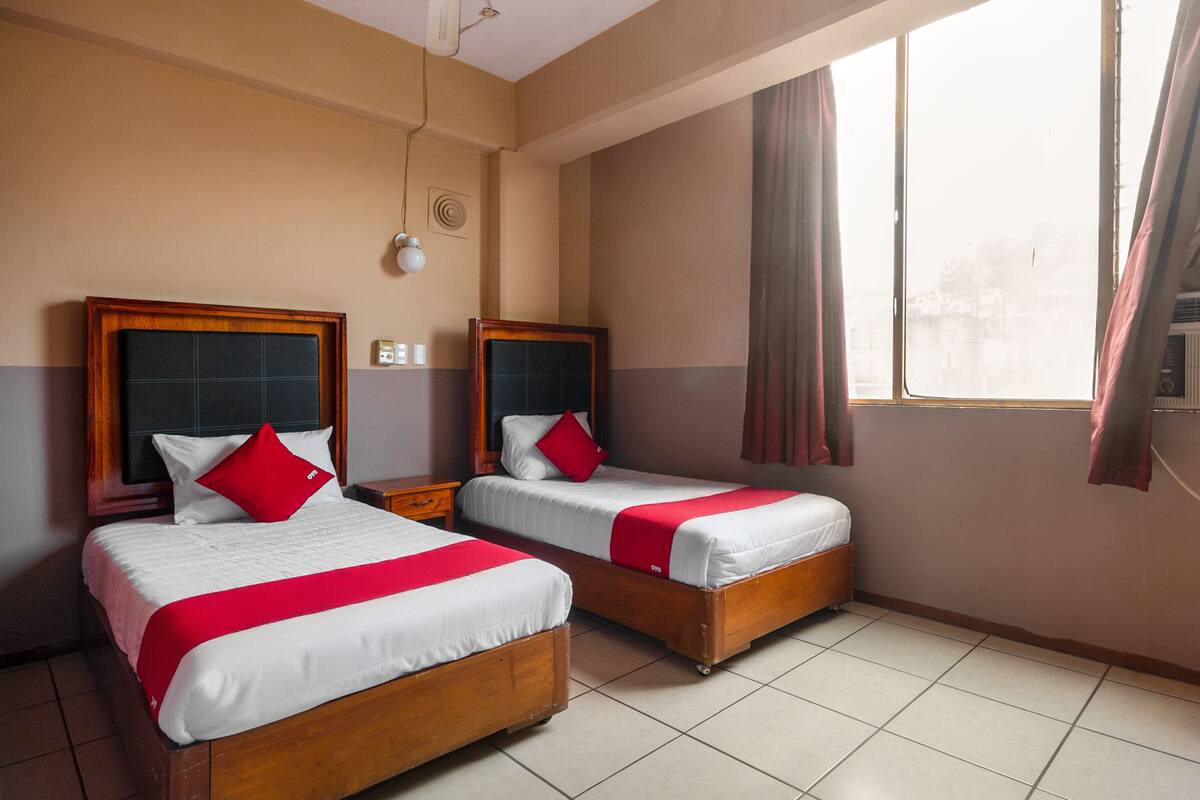 A hotel room with two wooden beds, each with white bedding and red pillows. There is a ceiling fan, a window with curtains, and a tiled floor.