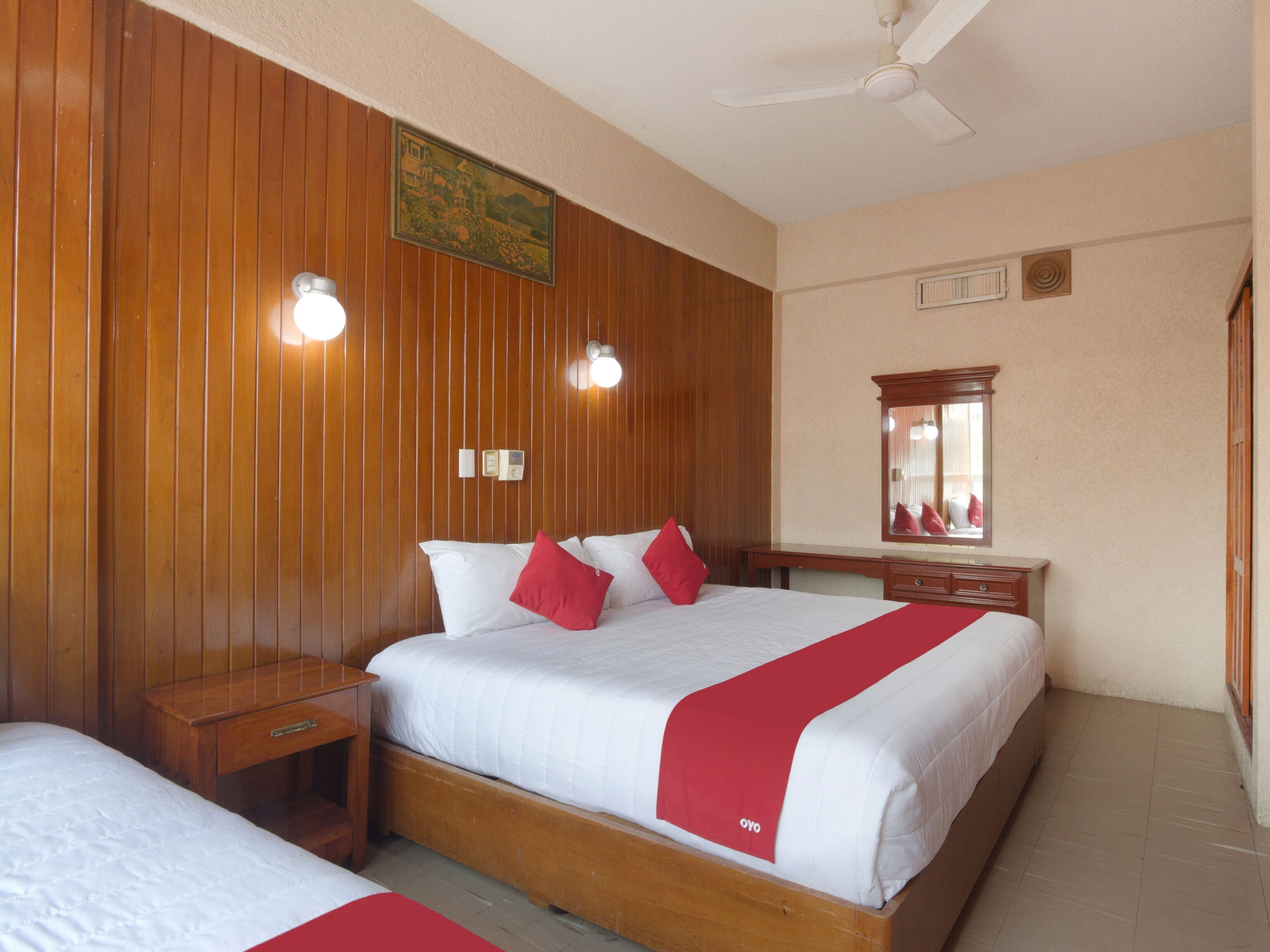 A hotel room with a wooden paneled wall, a bed with white bedding and red accents, a nightstand, a desk with a mirror, and a ceiling fan.