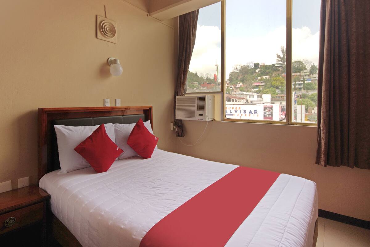 A hotel room with a bed, a nightstand, an air conditioning unit, a window with curtains, and a view of buildings outside.