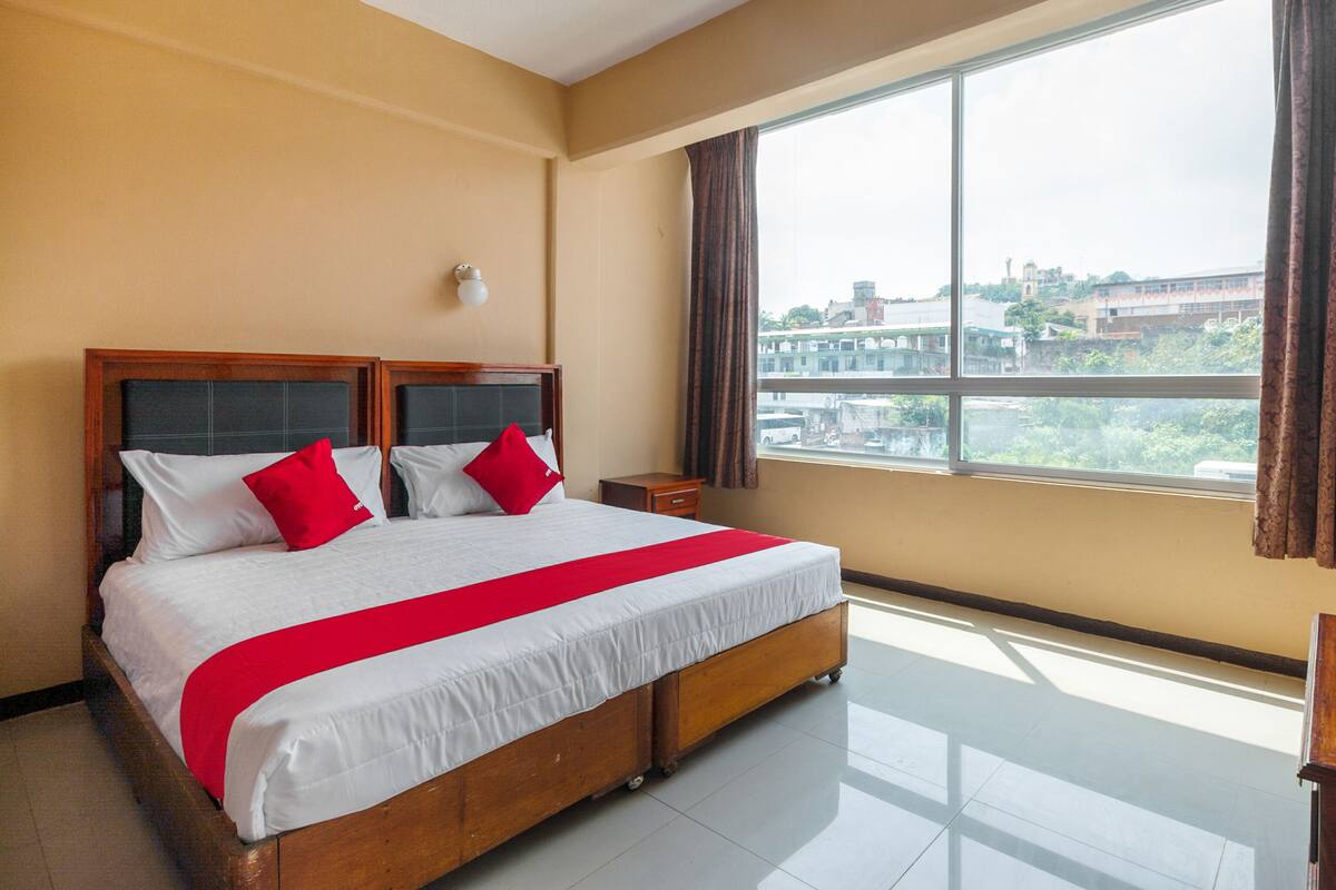 A hotel room with a large bed, a nightstand, a window with curtains, and a view of buildings outside.